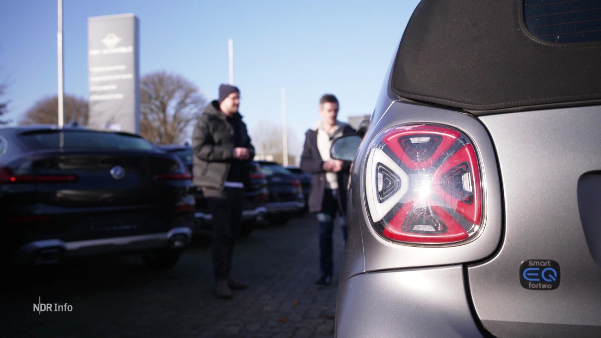 Rücklicht eines E-Autos im Vordergrund, im Hintergrund unscharf links daneben zwei Personen auf einem Parkplatz. | Screenshot