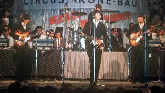 Die Beatles bei einem Live-Auftritt im Jahr 1966 im Circus Krone in München. Vor einem silbernen Vorhang stehen George Harrison, Paul McCartney und John Lennon mit Gitarren auf der Bühne. Ringo Starr sitzt am Schlagzeug. Alle tragen dunkle Anzüge.