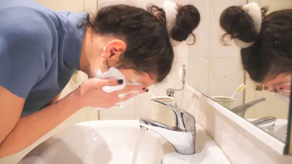 Eine Frau lehnt über einem Waschbecken und wäscht ihr Gesicht mit einem Schaum.