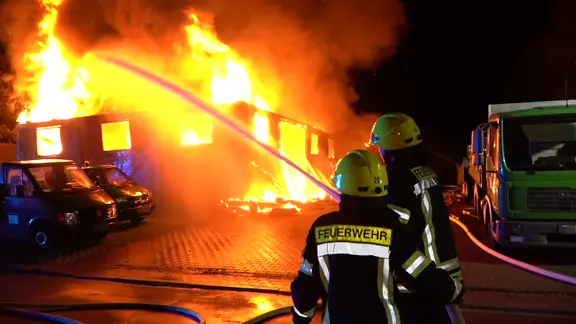 Feuerwehrleute löschen bei Nacht eine brennende Lagerhalle.