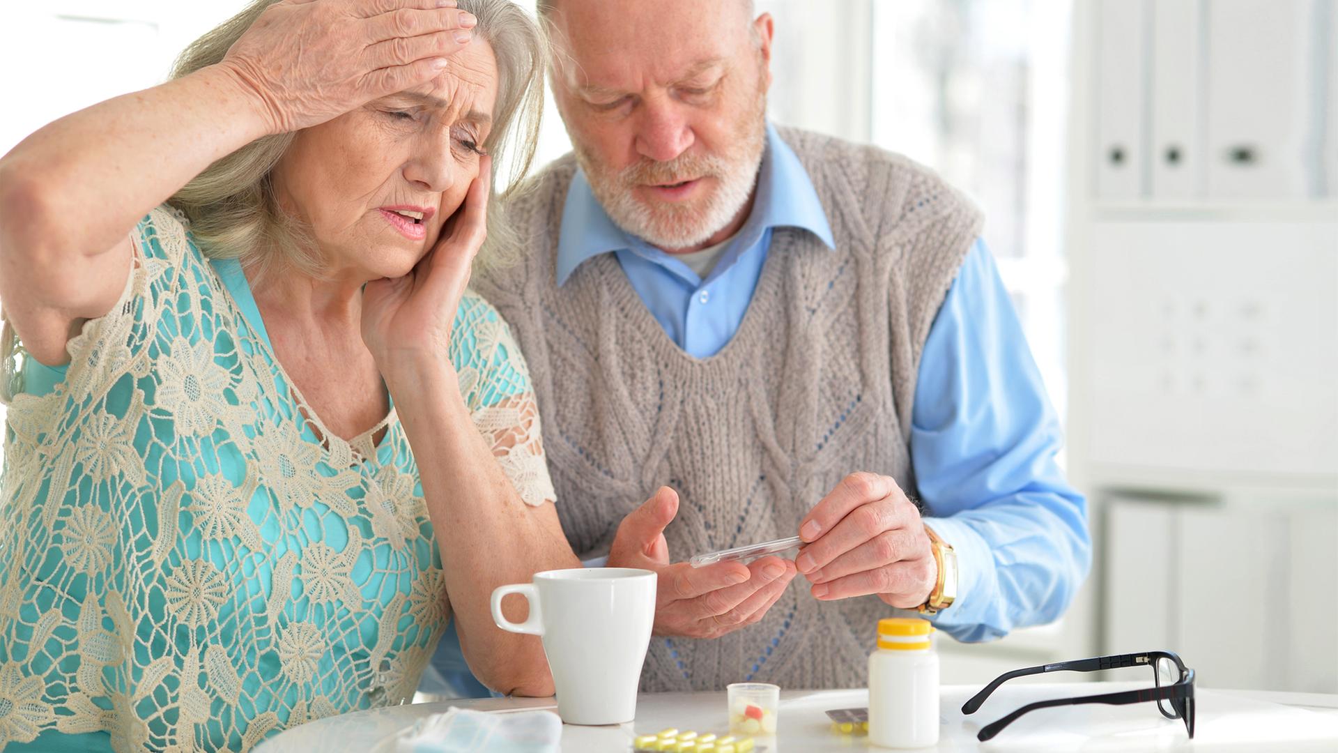 Seniorin fasst sich an die Schläfe, neben ihr Senior, der ein Thermometer betrachtet. | COLOURBOX, -