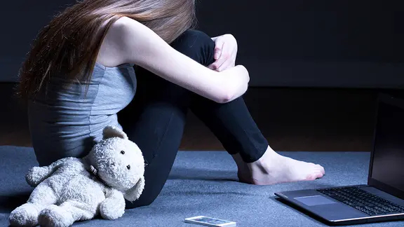 Mädchen mit Teddy kauert verzweifelt vor einem Laptop.
