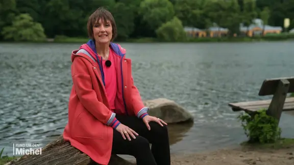 Theresa Pöhls moderiert die Sendung "Rund um den Michel" an einem See, der im Hintergrund zu sehen ist.