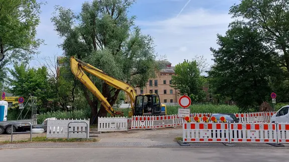 Auf dem Bild ist das Ende der Neuen Bleicherstraße zu sehen, an dem sich eine Baustelle befindet. Rot-weiße Absperrungen und ein Bagger sowie ein Lastanhänger stehen an dem Ufer des Seitenarmes der Warnow, bei dem die Fahrradbrücke gebaut werden soll. Die stelle befindet sich gegenüber dem Mühlendamm am Flussarm.