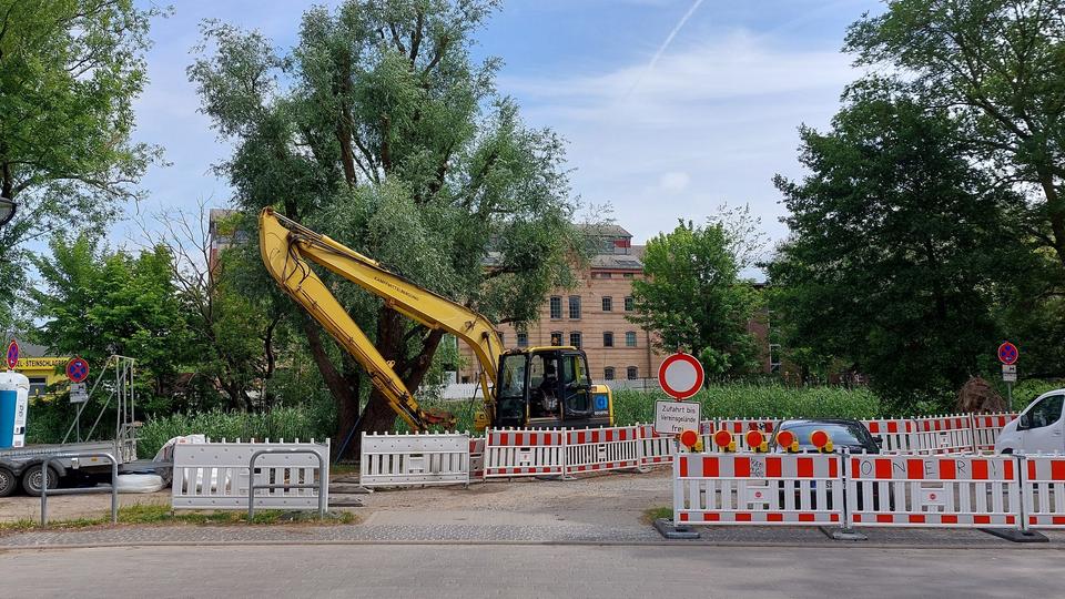 Auf dem Bild ist das Ende der Neuen Bleicherstraße zu sehen, an dem sich eine Baustelle befindet. Rot-weiße Absperrungen und ein Bagger sowie ein Lastanhänger stehen an dem Ufer des Seitenarmes der Warnow, bei dem die Fahrradbrücke gebaut werden soll. Die stelle befindet sich gegenüber dem Mühlendamm am Flussarm.
