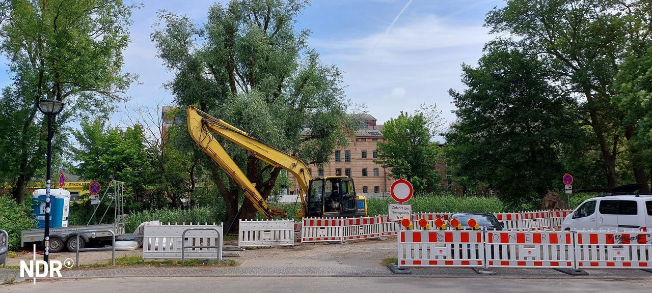 Auf dem Bild ist das Ende der Neuen Bleicherstraße zu sehen, an dem sich eine Baustelle befindet. Rot-weiße Absperrungen und ein Bagger sowie ein Lastanhänger stehen an dem Ufer des Seitenarmes der Warnow, bei dem die Fahrradbrücke gebaut werden soll. Die stelle befindet sich gegenüber dem Mühlendamm am Flussarm.