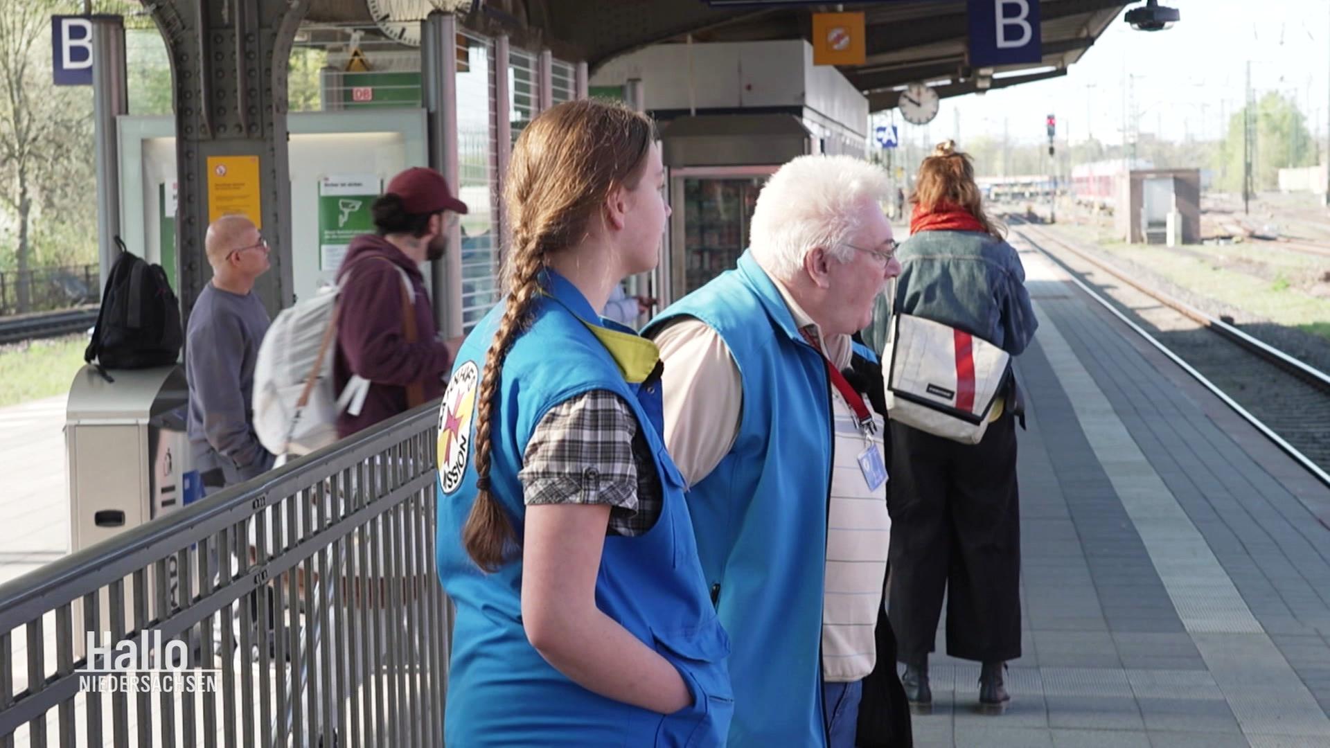 Zwei Mitarbeitende der Bahnhofsmission stehen am Bahnhof in Göttingen am Gleis. | Screenshot