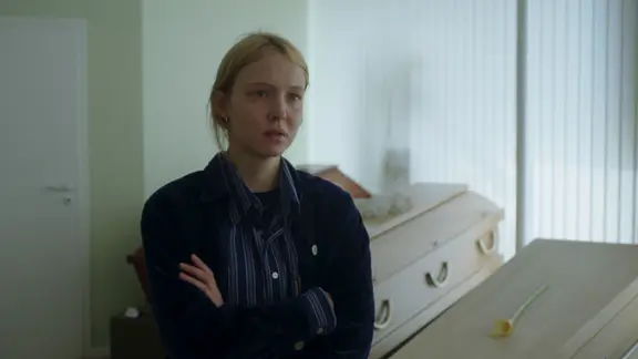 Eine Frau mit blonden Haaren steht mit verschränkten Armen und entsetztem Blick vor Holzsärgen.