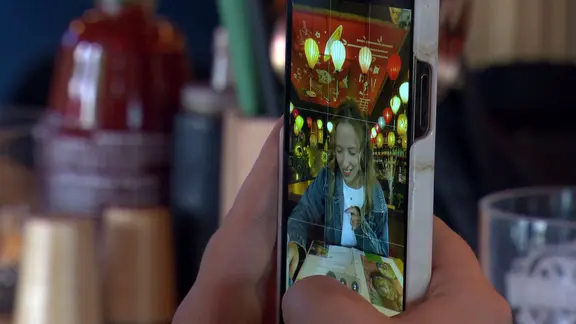 Hände halten ein Smartphone, mit dem eine Frau im Restaurant gefilmt wird.