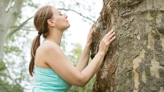 Eine Frau steht mit geschlossenen Augen an einem Baum, den sie mit ihren Händen berührt.