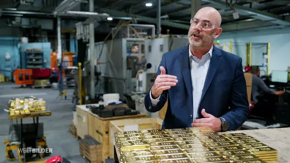 Ein Mann mit Schutzbrille steht vor einem Stapel mit Goldbarren.