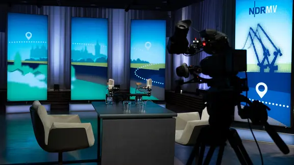 Ein Fernsehstudio mit vier hellen Sesseln um einen Tisch, auf dem mehrere Wassergläser stehen.