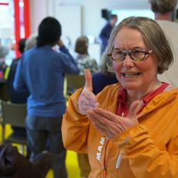 Eine Frau in orangefarbener Kapuzenjacke blickt in die Kamera und erklärt etwas in Gebärdensprache.
