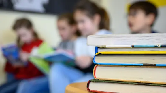 Kinder lesen in einer Grundschule, im Vordergrund ein Stapel Bücher