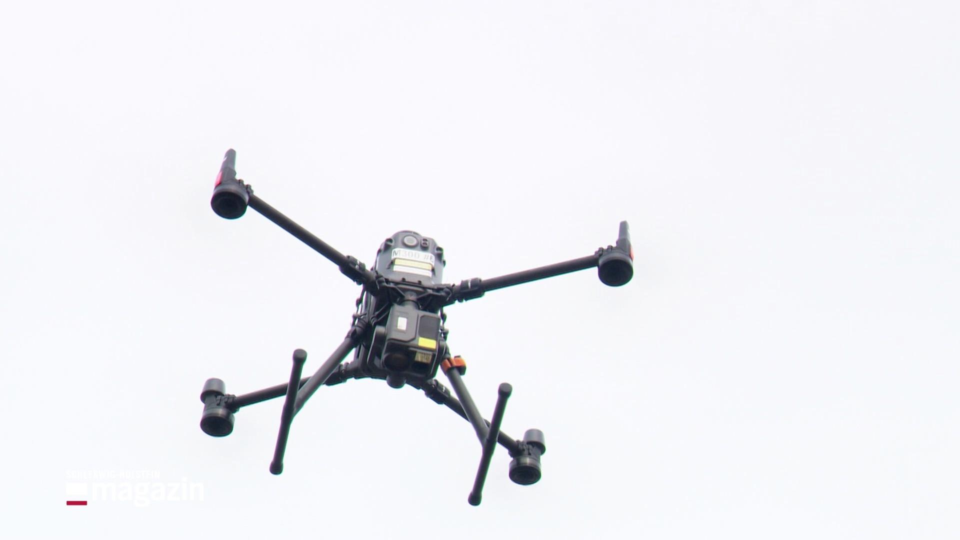 Eine schwarze Drohne mit vier Rotoren schwebt vor einem hellen, wolkigen Himmel. | Screenshot