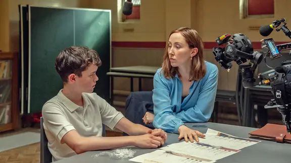 Ein Teenager-Junge mit dunklen kurzen Haaren sitzt in einem Verhör an einem Tisch mit einer Frau mit schulterlangen Haaren in einem großen Raum mit großem Tisch - Szene aus der "Netflix"-Serie "Adolescence" mit Schauspieler Owen Cooper