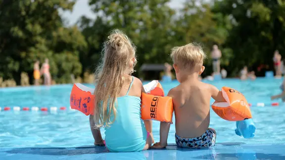 Zwei Kinder sitzen mit Schwimmflügeln am Rand eines Freibads.