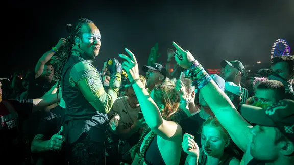 Ein Musiker von The Prodigy steht mitten im feiernden Publikum, das die Hände in die Höhe reckt und ausgelassen tanzt.