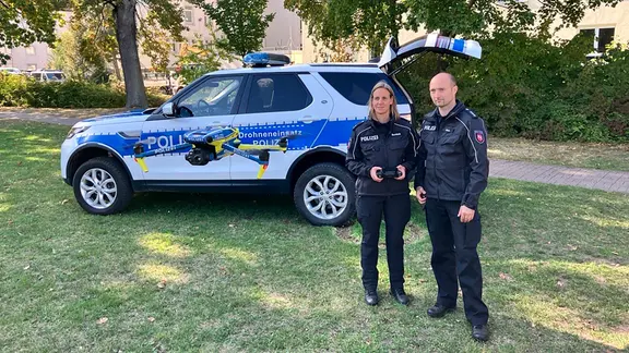 Zwei Polizisten stehen vor einem Polizeiauto und starten eine Drohne.