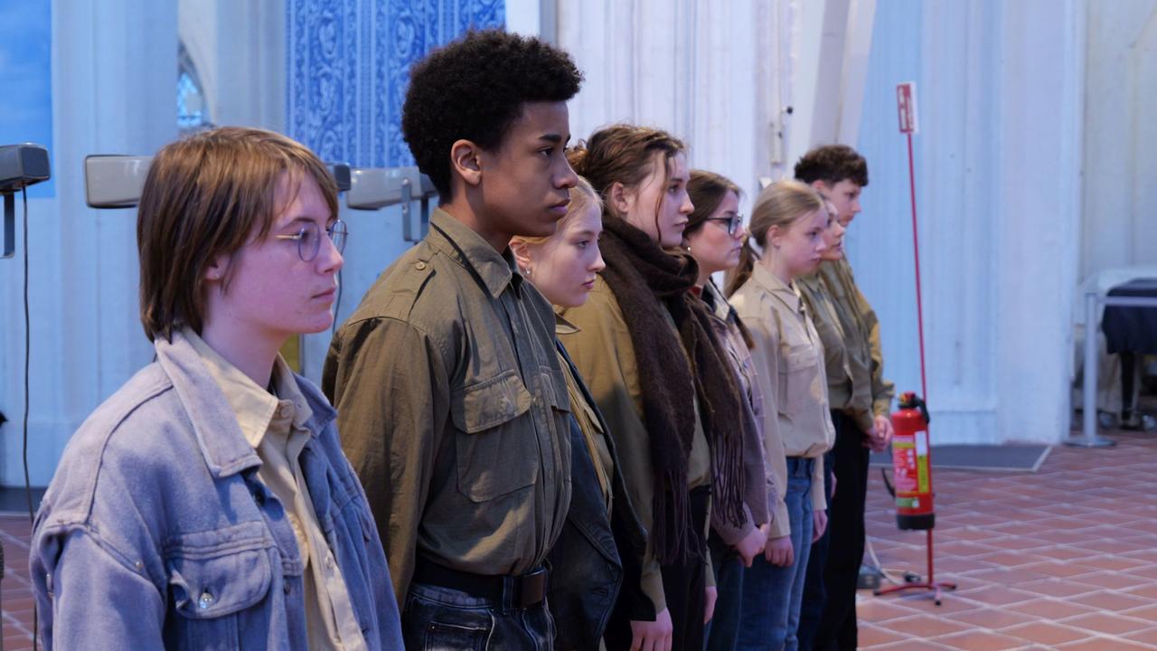 Acht Jugendliche stehen vor Holzhockern nebeneinander in einer Kirche.