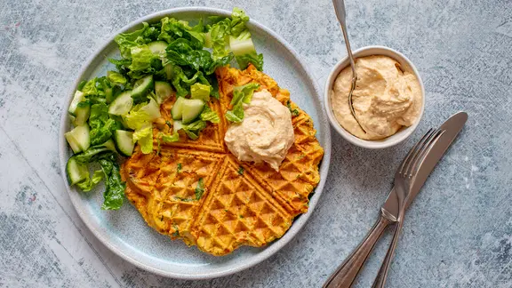 Ein Teller mit einer Waffel und einem grünen Salat, daneben ein Schälchen Hummus.