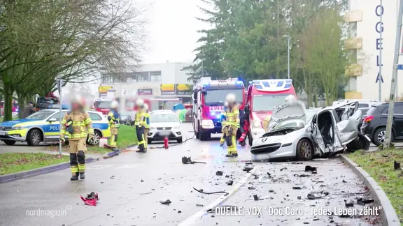 Bei einem schweren Autounfall steht ein komplett verbeulter PKW am Straßenrand einer Straße in einem Wohngebiet, Einsatzkräfte der Feuerwehr sichern die Unfallstelle.