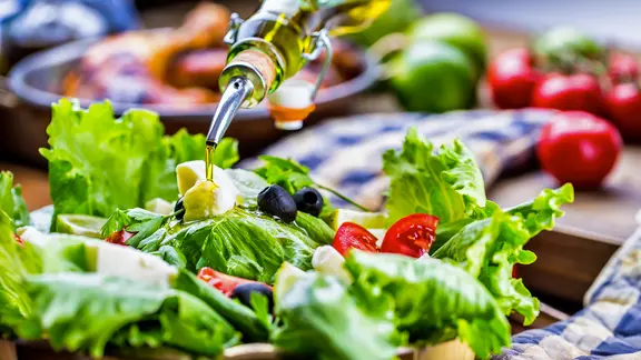 Ein Strahl Olivenöl ergießt sich über eine Schale mit Salat, Tomaten und Oliven