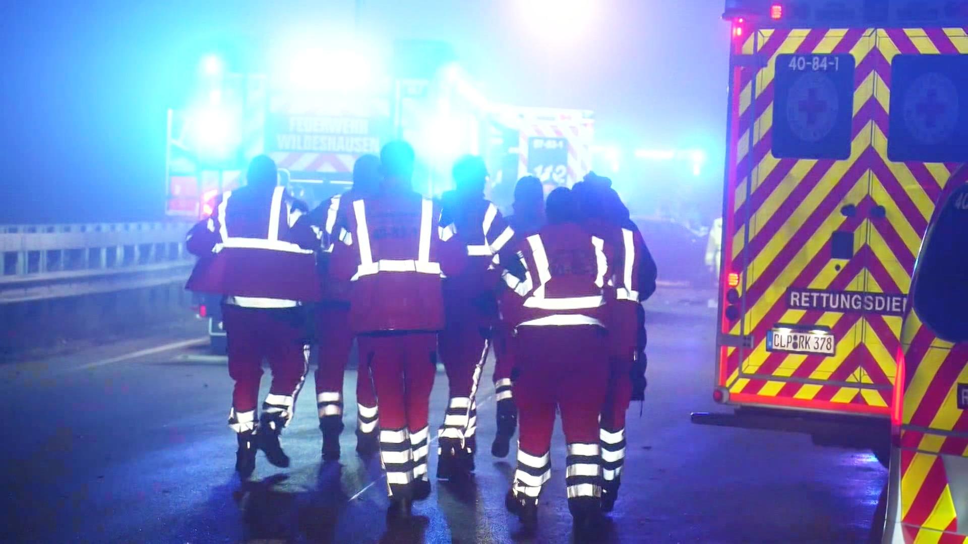 Mehrere Rettungskräfte in reflektierender Einsatzkleidung gehen in einer Gruppe auf einer nassen Straße zwischen Rettungsfahrzeugen mit eingeschaltetem Blaulicht, nachts bei Nebel. | Screenshot
