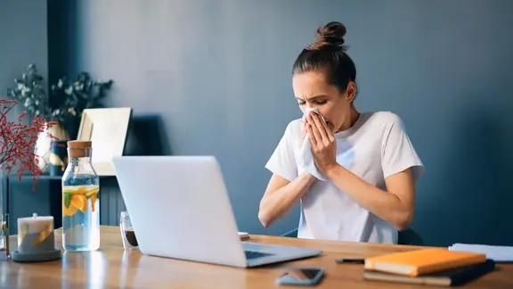 Eine junge Frau sitzt vor einem Laptop und schneuzt sich die Nase.