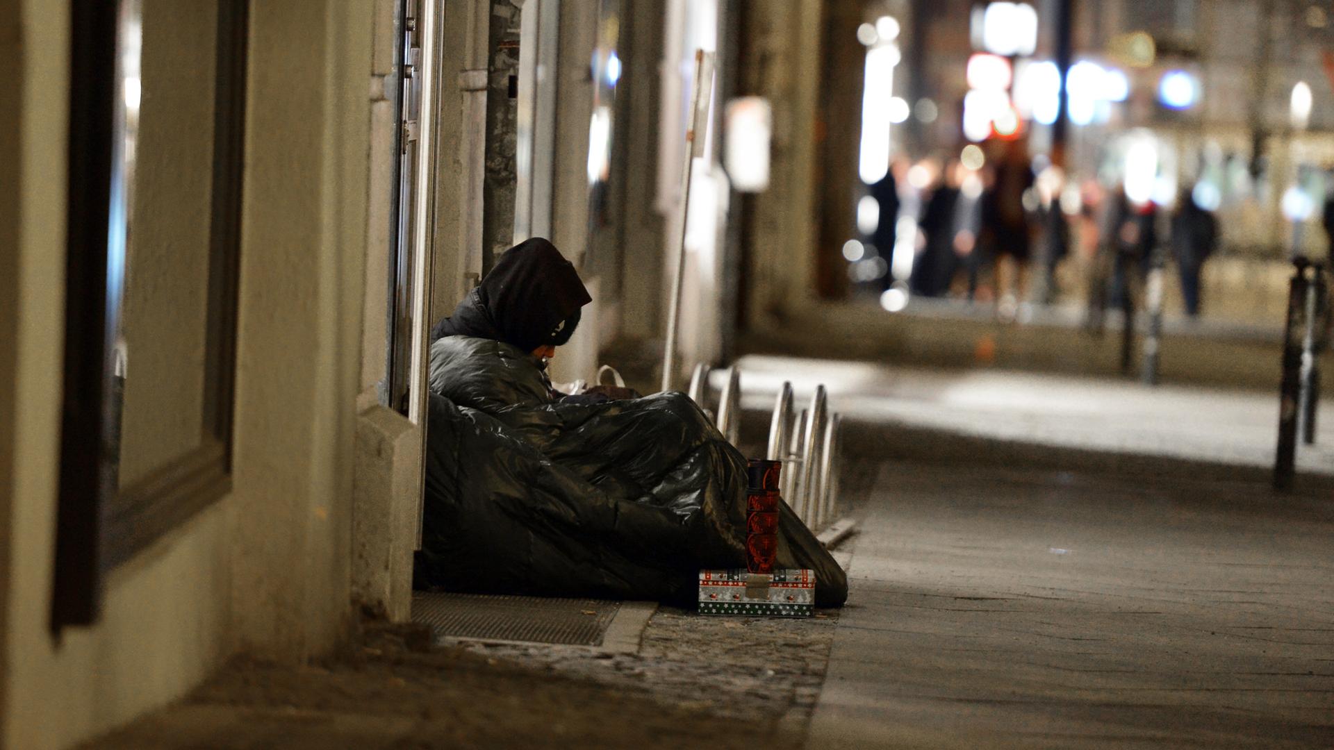Ein Obdachloser sitzt in einen Schlafsack gehüllt auf einem Gehweg | picture alliance / ZB | Jens Kalaene
