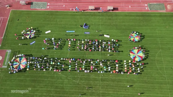 Menschen stehen auf einem Sportplatz und formen mit Regenschirmen einen Schriftzug.