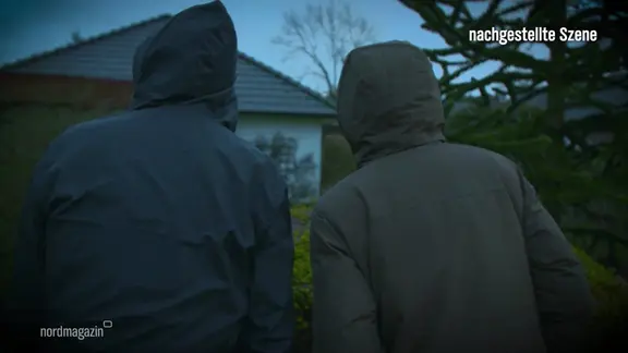 Zwei Männer mit Kaputze stehen in der Dämmerung vor einem Haus.