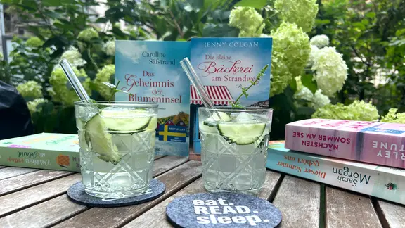 Mehrere aufgestellte hardcover-Bücher stehen auf einem Holztisch hinter Gläsern mit heller Füssigkeit (Cocktails)