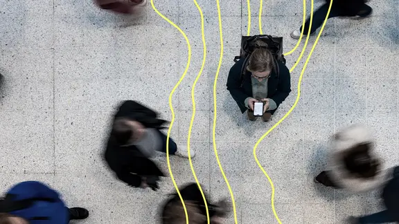 Eine Frau steht mit ihrem Handy in der Mitte während Passanten um sie herumgehen.