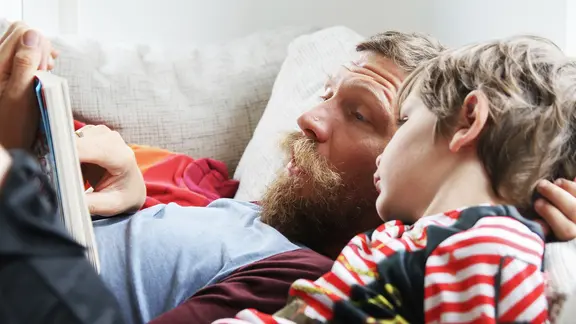 Ein Vater liest seinem Kind liegend auf dem Sofa vor.