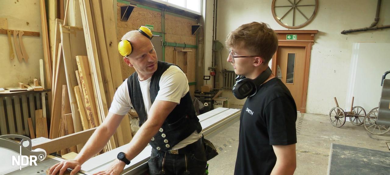 Ein Handwerker und sein Praktikant stehen in der Holzwerkstatt | Screenshot Ein Handwerker und sein Praktikant stehen in der Holzwerkstatt