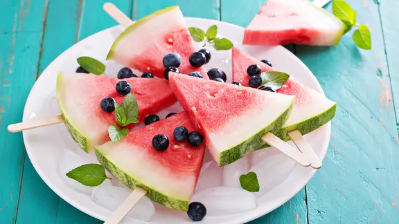 Ein Teller mit Wassermelonen-Stücken am Stiel. Darüber liegen Blaubeeren verteilt.