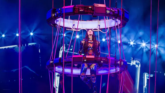 Eine Frau in einem futuristischen Kostüm, umgeben von zwei großen Ringen, die von der Decke hängen, steht auf der Bühne und blickt nach oben