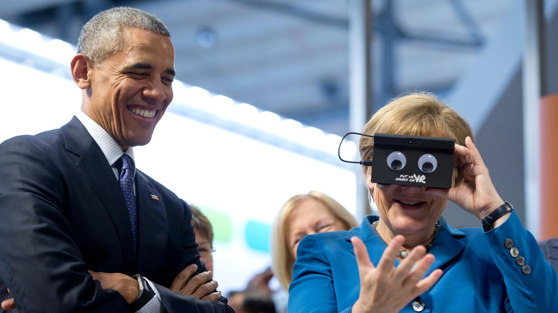Bei der Hannover Messe 2016 stehen Barack Obama und Angela Merken nebeneinander. Merkel testet eine VR-Brille. | picture alliance/AP Photo | Carolyn Kaster