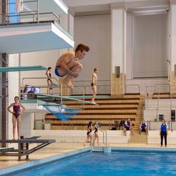 Ein junger Turmspringer springt in der Hocke vom Sprungbrett in ein Schwimmbecken. Mehrere Kinder und Jugendliche stehen am Beckenrand oder auf anderen Sprungplattformen. Im Hintergrund sind leere Tribünen zu sehen.