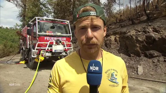 Reporter Tino Nowitzki vor Ort im Harz, im Hintergrund ein Feuerwehrauto. 