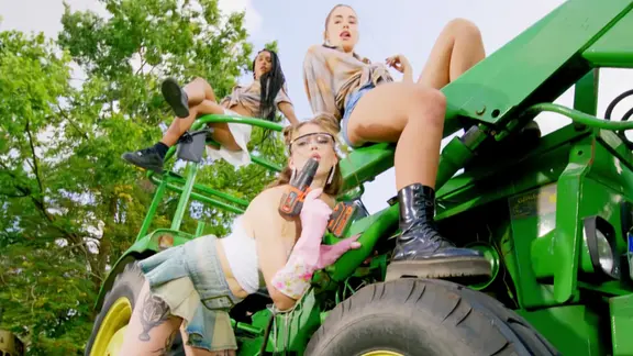 Drei leicht begleidete Frauen räkeln sich auf einem Traktor.