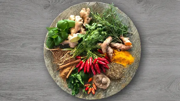 Ein schön angerichteter Teller mit Kurkuma, Chili, Ingwer und verschiedenen weiteren Gewürzen steht auf einer Holzplatte.