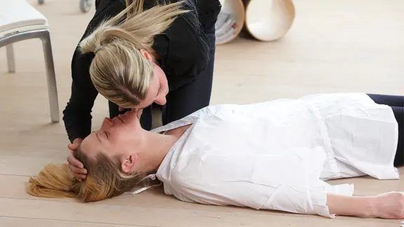 Eine Frau horcht an dem Atem einer Frau, die am Boden liegt.