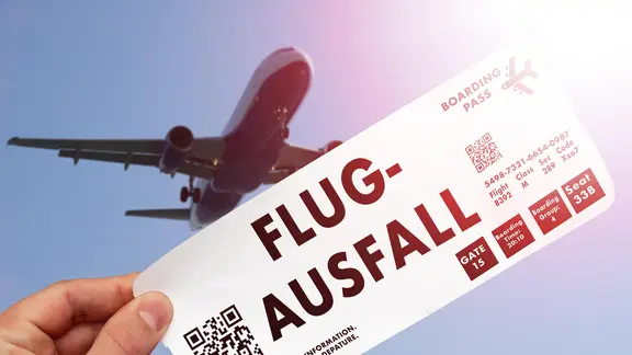 Ein Flugticket mit der Aufschrift "Flugausfall" wird vor einem startenden Flugzeug gehalten.