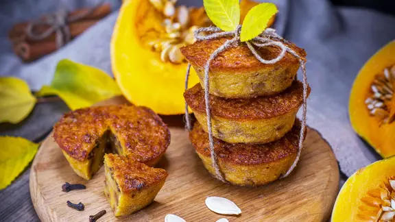 Ein kleiner Stapel mit Kürbis-Muffins auf einem Holzbrett, dahinter ist ein Stück Kürbis zu sehen.