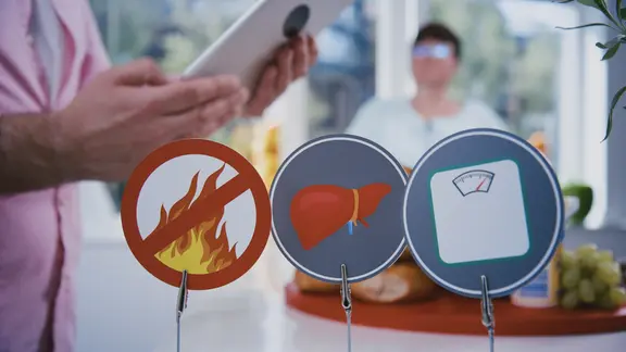 Drei Symbolschilder auf einem Tisch: links durchgestrichene Flamme, in der Mitte eine Leber und rechts eine Waage.