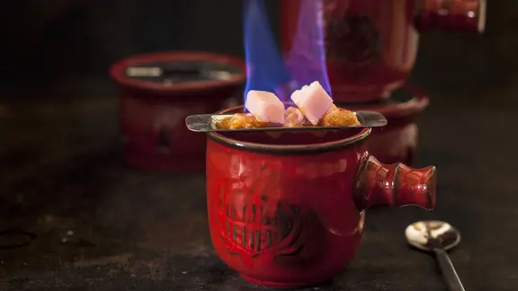 Statt gleich einen ganzen Topf Feuerzangenbowle zuzubereiten, kann man auch einzelne Becher mit dem Heißgetränk befüllen. | Imago/Westend61 Eine Mini-Feuerzangenbowle brennt in einem dunklen Raum.