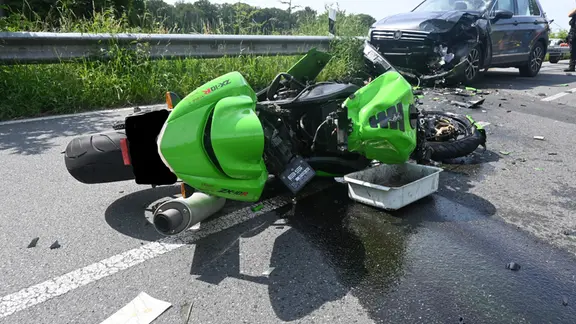 Ein grünes Motorrad liegt schwer beschädigt auf einer Straße vor einem Auto mit Frontalschaden 