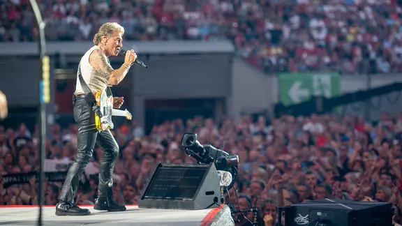 Ein Mann steht vor vielen Menschen in einem Stadion auf einer Bühne und singt.